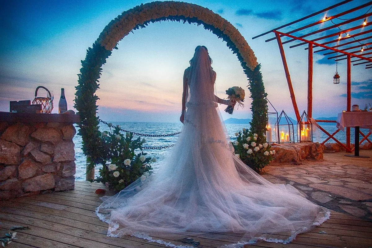 A bride beneath the floral arch on the pier at sunset, seen from behind.